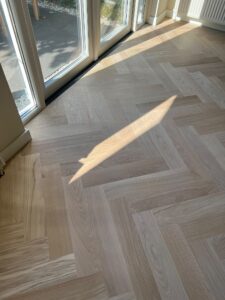 Zonlicht stroomt door de glazen deuren naar binnen en valt op een lichte houten visgraatvloer, waardoor er een scherpe, geometrische schaduw over het patroonrijke oppervlak valt.