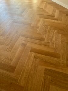 Lichtbruine houten vloer met een visgraatpatroon, gefotografeerd in natuurlijk daglicht. Het hout heeft een gladde afwerking en de verschillende bruintinten laten het geometrische patroon goed tot zijn recht komen.