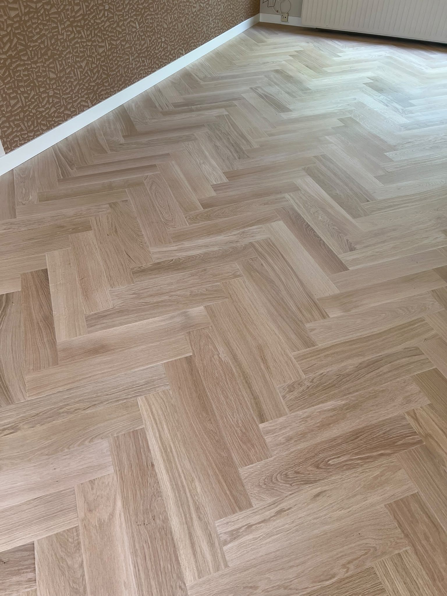 Een lichte houten vloer in visgraatpatroon, grenzend aan een bruine muur met patroon en een witte plint, terwijl het zonlicht rechtsboven naar binnen stroomt.