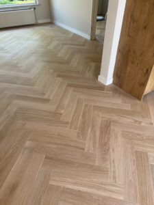 Een vloer met een visgraatpatroon van licht hout in een lichte, lege kamer met crèmekleurige muren, een raam met daaronder een radiator en een open deuropening die naar een andere kamer leidt.