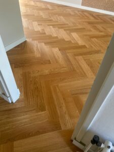 Een kamer met een houten vloer in visgraatpatroon, gezien vanuit een open deuropening. In de rechteronderhoek zijn een klein stukje van een witte muur en een huisdierhekje te zien.