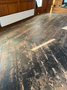 Het zonlicht schijnt op een grote, versleten houten vloer met zichtbare krassen en slijtsporen. Aan één kant van de kamer bevinden zich met hout beklede muren en een witte radiator.