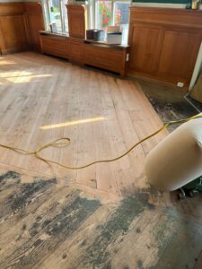 De houten vloer is gedeeltelijk geschuurd; sommige delen zijn licht en glad, andere donker en versleten. In een zonovergoten kamer met houten lambrisering en grote ramen zijn een geel verlengsnoer en een schuurmachine te zien.