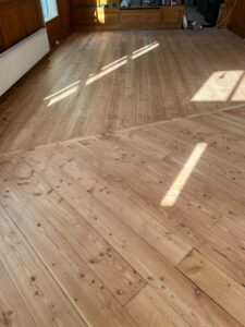Zonlicht stroomt binnen op een houten vloer met brede planken in een grotendeels lege kamer. De vloerplanken komen schuin samen, waardoor er een duidelijke scheidingslijn en een patroon in het hout ontstaat. De muren zijn bekleed met houten lambrisering en er staat een witte radiator.