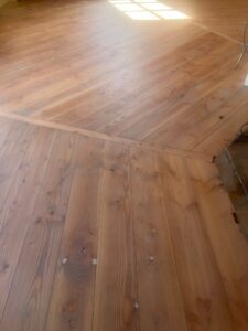Het zonlicht schijnt op een schone, afgewerkte houten vloer met diagonale plankpatronen die onder een hoek samenkomen; rechts zijn enkele draden en een onafgewerkte rand zichtbaar.