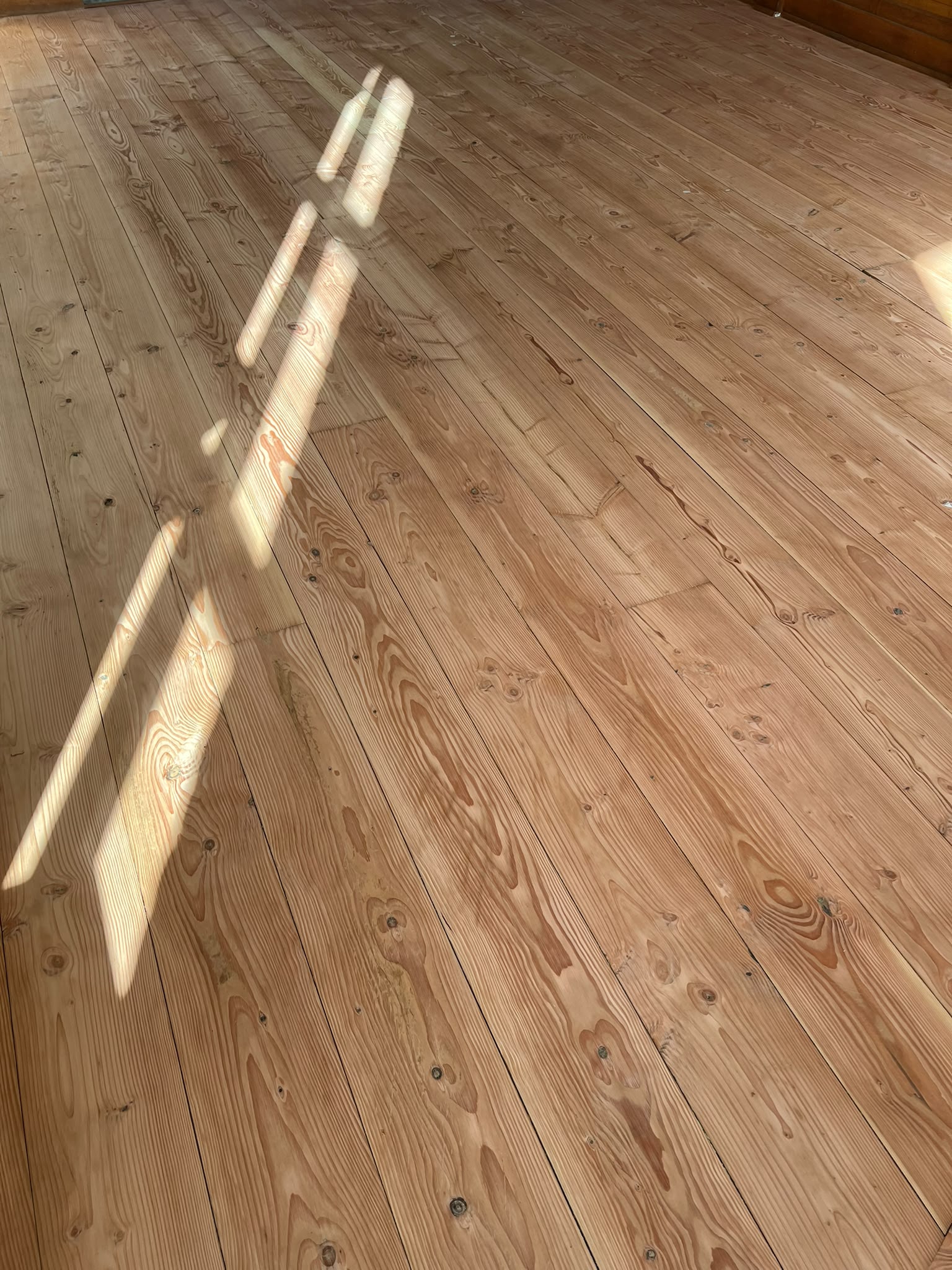 Het zonlicht stroomt door een raam naar binnen en werpt heldere rechthoekige patronen op een schone, gepolijste houten vloer met zichtbare natuurlijke nerven en knoesten.
