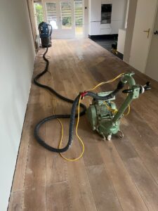 Op de houten vloer van een moderne woning staan een groene vloerschuurmachine en een stofzuiger, terwijl kabels en slangen zich door de gang uitstrekken in de richting van een zonovergoten deuropening.