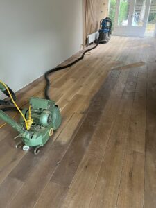 Op een houten vloer, die gedeeltelijk geschuurd is, staat een vloerschuurmachine. De kamer is leeg; op de achtergrond staat een stofzuiger en door de grote ramen valt licht naar binnen.