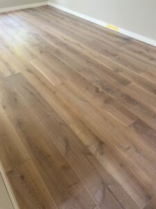 Een lichtbruine houten vloer met zichtbare houtnerf in een lege kamer, witte plinten en beige muren. Bij de muur aan de andere kant is een klein geel voorwerp te zien.