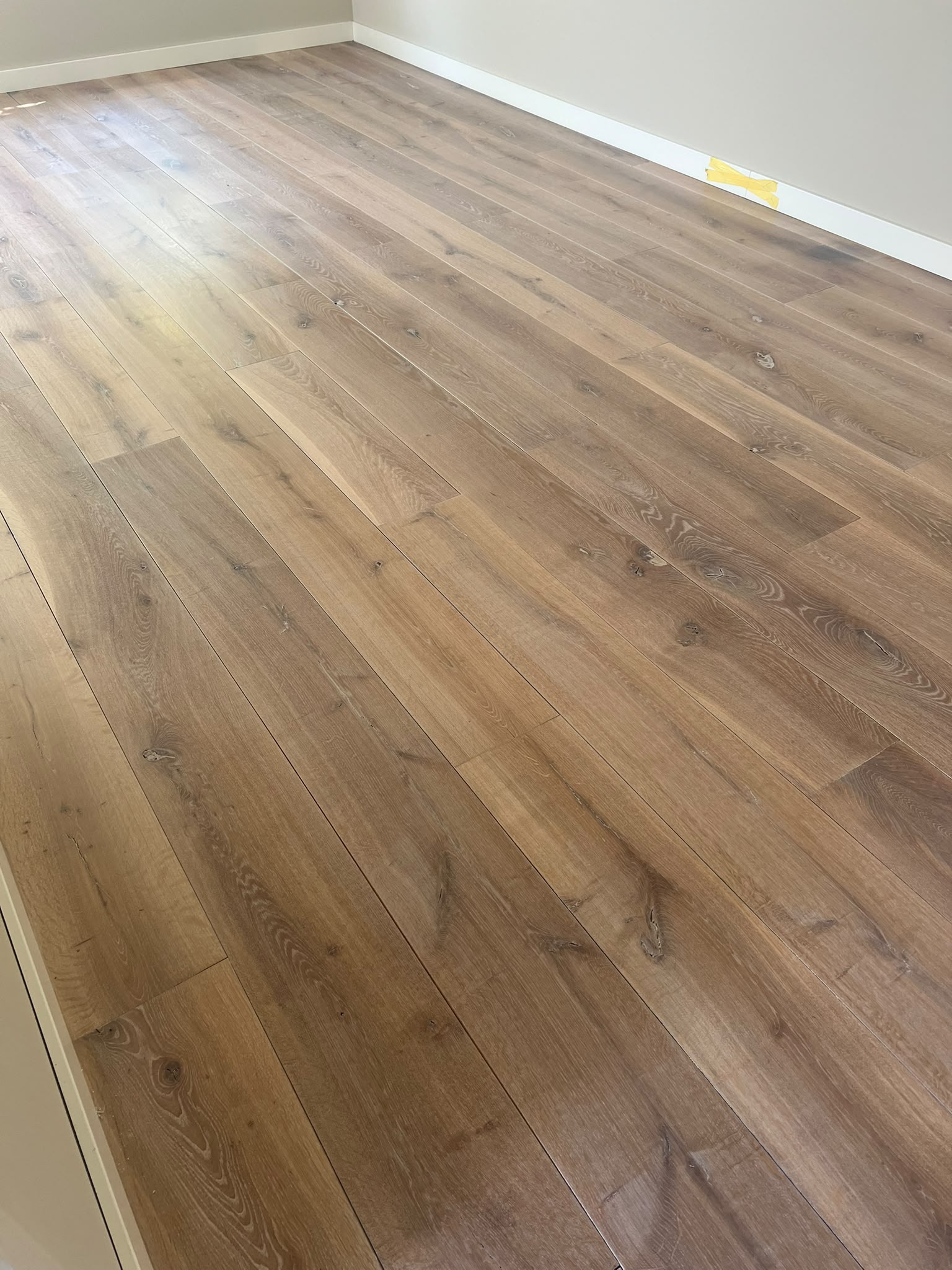 Een kamer met een lichtbruine houten vloer, witte plinten en beige muren. Op de vloer, vlakbij de achterste muur, ligt een klein geel voorwerp. De kamer ziet er leeg en goed verlicht uit.